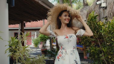 Genç ve güzel gülümseyen Avrupalı esmer kız arka planda tropikal çardağı olan hasır bir şapkayla arka bahçede yürüyor.