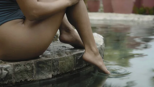 The legs of a young and beautiful European girl in a swimming suit close-up sitting near swimming pool with barefoot wet legs touching her legs with her hands
