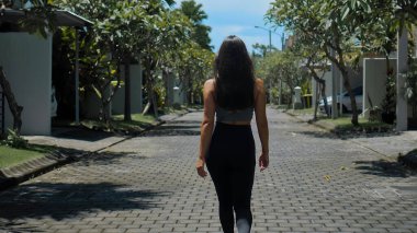 Genç Avrupalı esmer bir spor kızı, arka planda yeşil ağaçlarla boş bir şehir caddesinde yürüyor.