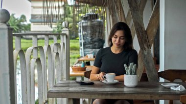 Genç Avrupalı bir kız halka açık bir yerde bir fincan kahveyle ahşap bir masada oturuyor. 