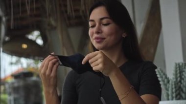 Genç Avrupalı esmer bir kız halka açık bir masada oturuyor ve yüzüne siyah koruyucu bir maske takıyor.