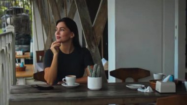 Genç Avrupalı bir kız halka açık bir yerde elinde bir fincan kahveyle masada oturuyor ve yanında da siyah koruyucu bir maske var.