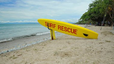 Plajda sarı sörf tahtası ve Bali adasındaki bir sahilde acil durum mesajı.