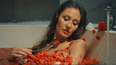 Genç Avrupalı uzun saçlı esmer kız banyoda, spa prosedüründe kırmızı çiçeklerle yatıyor.