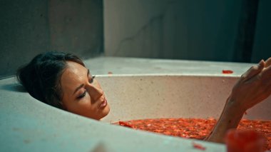 Genç Avrupalı uzun saçlı esmer kız banyoda, spa prosedüründe kırmızı çiçeklerle yatıyor.