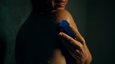 Genç Avrupalı uzun saçlı esmer kız elinde mavi sabunla koyu bir duşta duruyor.