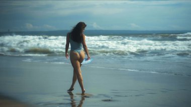 Bikinili genç, seksi Avrupalı bir kız mavi dalgalarla deniz kenarında siyah kumlarda yürüyor.