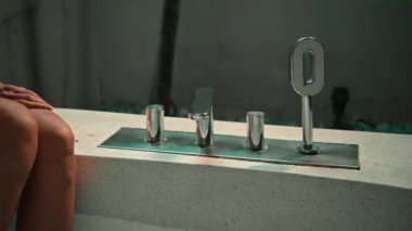 Küvette oturan genç Avrupalı bir kız banyoda elleriyle su dolu bir musluk açıyor.