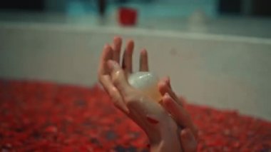 Genç Avrupalı uzun saçlı esmer kız banyoda, spa prosedüründe kırmızı çiçeklerle gül şeklinde sabunla yatıyor.