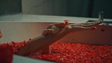 Genç Avrupalı uzun saçlı esmer kız banyoda, spa prosedüründe kırmızı çiçeklerle gül şeklinde sabunla yatıyor.