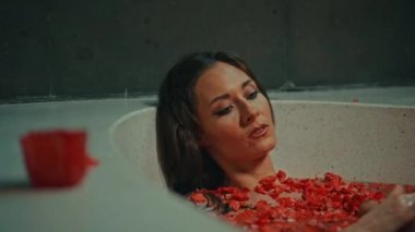 Genç Avrupalı uzun saçlı esmer kız banyoda, spa prosedüründe kırmızı çiçeklerle gül şeklinde sabunla yatıyor.