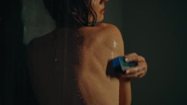 Genç Avrupalı uzun saçlı esmer kız elinde mavi sabunla koyu bir duşta duruyor.