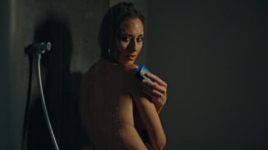 Genç Avrupalı uzun saçlı esmer kız elinde mavi sabunla koyu bir duşta duruyor.