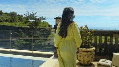 Arka bahçede yeşil bahçeli, mavi temiz su havuzlu ve mavi gökyüzü güzel, sarı elbiseli, uzun saçlı genç bir kız yürüyor.