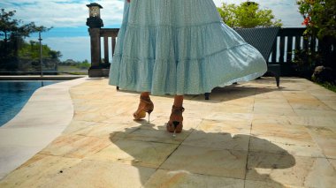 Bahçede yeşil bir bahçeyle yürüyen genç bir kız, mavi temiz su ile havuz ve güzel mavi bir elbise içinde mavi gökyüzü. 