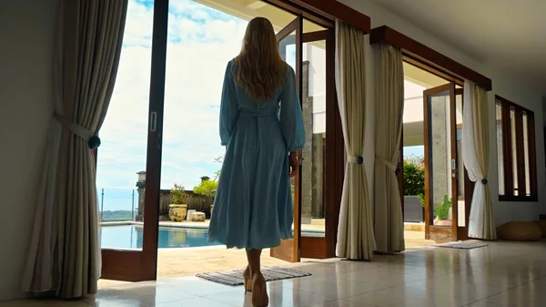 Mavi elbiseli genç kız avluya bakan geniş kapıları ve mavi gökyüzü ve bulutları olan tropiklerde büyük bir havuzu olan evi terk ediyor.