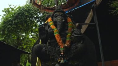 Ganesha 'nın eski taş heykeli, boynunda sarı bir çiçek çelengi olan yosunlarla kaplıydı. Arka planda yeşil bitkilerle sokakta duruyordu.