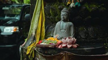 Nilüfer çiçekli eski bir Budist heykeli. Boynunda sarı bir çiçek çelengi var. Arka planda yeşil bitkiler var.