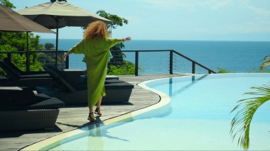 Uzun, kırmızı kıvrımlı, yeşil bahçeli, mavi temiz su havuzlu ve güzel yeşil elbiseli, mavi gökyüzü olan genç, saçları olan bir kız.