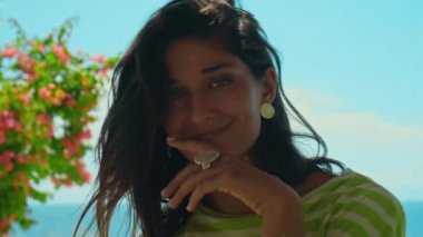 Genç Avrupalı esmer bir kız arka bahçede oturur ve gülümseyerek elleriyle yüzüne dokunur. Arka planda yeşil bitkiler vardır.