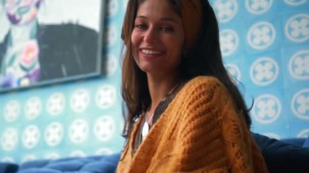 一位年轻貌美的欧洲姑娘 长发黑发 坐在沙发地板上的房间里 凝视着摄像机 微笑着触摸着脖子上的一条黄色围巾 — 图库视频影像