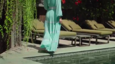 Genç Avrupalı uzun saçlı kız yeşil bahçeli bahçede yürüyor, mavi temiz su havuzlu güzel mavi elbisesi var. 