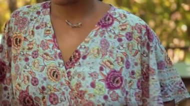 Uzun saçlı genç bir esmer kız nazikçe boynunda asılı olan kolyeye dokunur, arka bahçede durur.