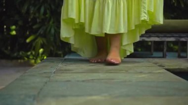 Genç Avrupalı bir kızın bacakları, arka bahçedeki çimlerin üzerinde yeşil bir bahçe ve güzel, sarı bir elbiseyle yürüyor.