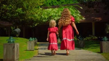 Anne ve kızı arka bahçede yeşil bir bahçe ve güzel kırmızı elbiseli çimlerle yürüyor, sırtını dönüyor.