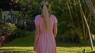Bahçede yeşil bahçesi ve güzel pembe elbisesi olan çimleriyle dolaşan genç bir Avrupalı kız arkasını dönüyor.