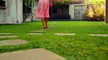 Bahçede yeşil bahçesi ve güzel pembe elbisesi olan çimleriyle dolaşan genç bir Avrupalı kız arkasını dönüyor.