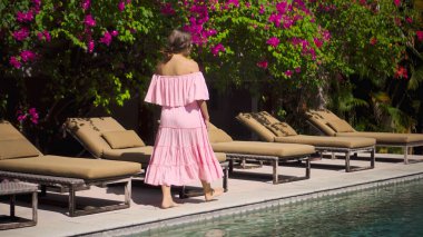 Genç Avrupalı uzun saçlı kız bahçesinde yeşil bir bahçeyle yürüyor, mavi temiz suyla dolu bir havuz güzel pembe elbisesi var. 