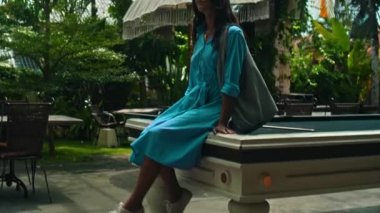 Mavi elbiseli genç bir Avrupalı kız ve elinde bir bayan çantasıyla tropik bölgelerde yeşil bir parkta yürüyor.