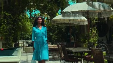 Genç ve güzel Avrupalı bir kız uzun saçlı esmer mavi bir elbise içinde yeşil bitkiler ve beyaz şemsiyelerle caddede yürüyor.