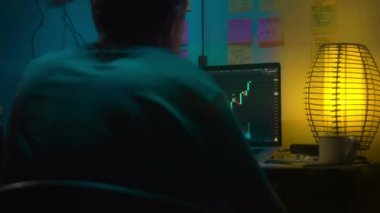Avrupalı bir kripto tüccarı karanlık bir odada, elinde neon ışığı olan bir dizüstü bilgisayarın arkasında oturur, kripto döviz değişimindeki fiyat hareketlerinin grafiklerini gösteren bir ekrana bakar. Bir sigara içiyor. Bulanık arkaplan