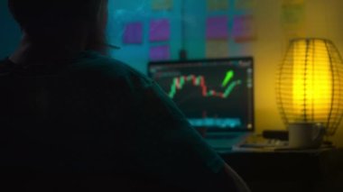 Avrupalı bir kripto tüccarı karanlık bir odada, elinde neon ışığı olan bir dizüstü bilgisayarın arkasında oturur, kripto döviz değişimindeki fiyat hareketlerinin grafiklerini gösteren bir ekrana bakar. Bir sigara içiyor. Bulanık arkaplan