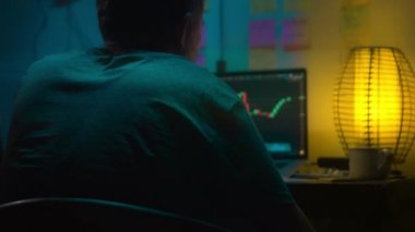 Avrupalı bir kripto tüccarı karanlık bir odada, elinde neon ışığı olan bir dizüstü bilgisayarın arkasında oturur, kripto döviz değişimindeki fiyat hareketlerinin grafiklerini gösteren bir ekrana bakar. Bir sigara içiyor. Bulanık arkaplan