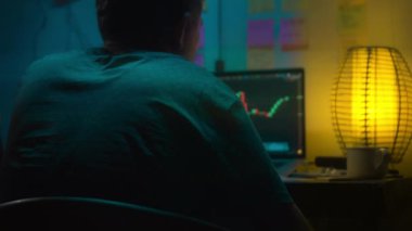 Avrupalı bir kripto tüccarı karanlık bir odada, elinde neon ışığı olan bir dizüstü bilgisayarın arkasında oturur, kripto döviz değişimindeki fiyat hareketlerinin grafiklerini gösteren bir ekrana bakar. Bir sigara içiyor. Bulanık arkaplan