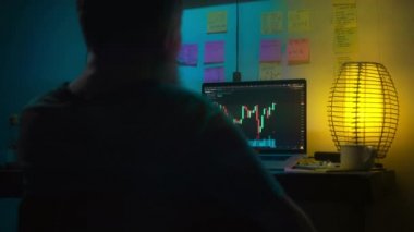 Avrupalı bir kripto tüccarı karanlık bir odada, elinde neon ışığı olan bir dizüstü bilgisayarın arkasında oturur, kripto döviz değişimindeki fiyat hareketlerinin grafiklerini gösteren bir ekrana bakar. Bir sigara içiyor. Bulanık arkaplan