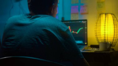 Avrupalı bir kripto tüccarı karanlık bir odada, elinde neon ışığı olan bir dizüstü bilgisayarın arkasında oturur, kripto döviz değişimindeki fiyat hareketlerinin grafiklerini gösteren bir ekrana bakar. Bir sigara içiyor. Bulanık arkaplan