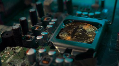 Karanlıkta mavi ışığı olan elektronik bileşenleri olan bir ana kartta merkezi işlemci yerine parlayan ve yansıtan altın bir Bitcoin. Bitcoin madenciliği.