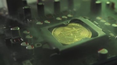 Karanlıkta ışık ve duman olan elektronik bileşenleri olan ana kartta merkezi bir işlemci yerine parlayan ve yansıtan altın bir Bitcoin. Bitcoin Madencilik