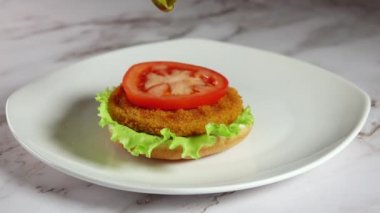 White plate on a marble table. Two slices of pickled cucumber fall on half a bun with a salad, a fish cutlet and a slice of tomato. Step 4 of 4. Slow motion