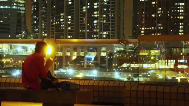 香港や高層ビルの風の夜 寂しい男がベンチに座ってスマホを見ている — ストック動画