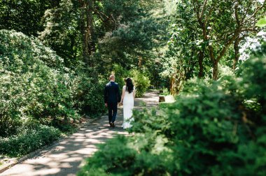Gelin ve damat yaz parkında, ormanda yürüyor. Balayı için düz yolda koşan yeni evli bir çift. Arka görünüm.