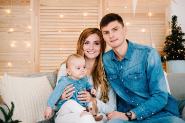 Baba, anne küçük kızını Noel ağacının yanındaki kanepede oturtuyor. Mutlu yıllar ve mutlu Noeller. Noel dekore edilmiş iç mekan. Aile kış tatili kavramı. Kapat..