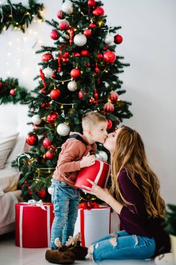 Anne oğluna Noel ağacının yanında bir hediye verir. Mutlu yıllar ve mutlu Noeller. Noel dekore edilmiş iç mekan. Aile tatili kavramı.