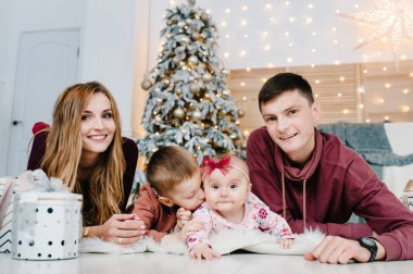 Baba ve anne, Noel ağacının yanında yerde yatan hediyelerle küçük oğlunu ve kızını kucaklıyor. Mutlu yıllar ve mutlu Noeller. Noel dekore edilmiş iç mekan. Aile tatili kavramı.