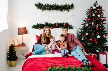 Baba ve anne, Noel ağacının yanındaki yatak odasında küçük oğlana ve kızına sarılıyor. Mutlu yıllar ve mutlu Noeller. Noel dekore edilmiş iç mekan. Aile tatili kavramı. Kapat..