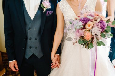 Gelin ve damat, bir düğün buketiyle el ele tutuşuyorlar ve evin içinde nikah töreninde duruyorlar..